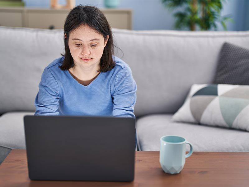 A person with Down Syndrome is sitting at a coffee table with a laptop open in front of them, looking down at the screen.