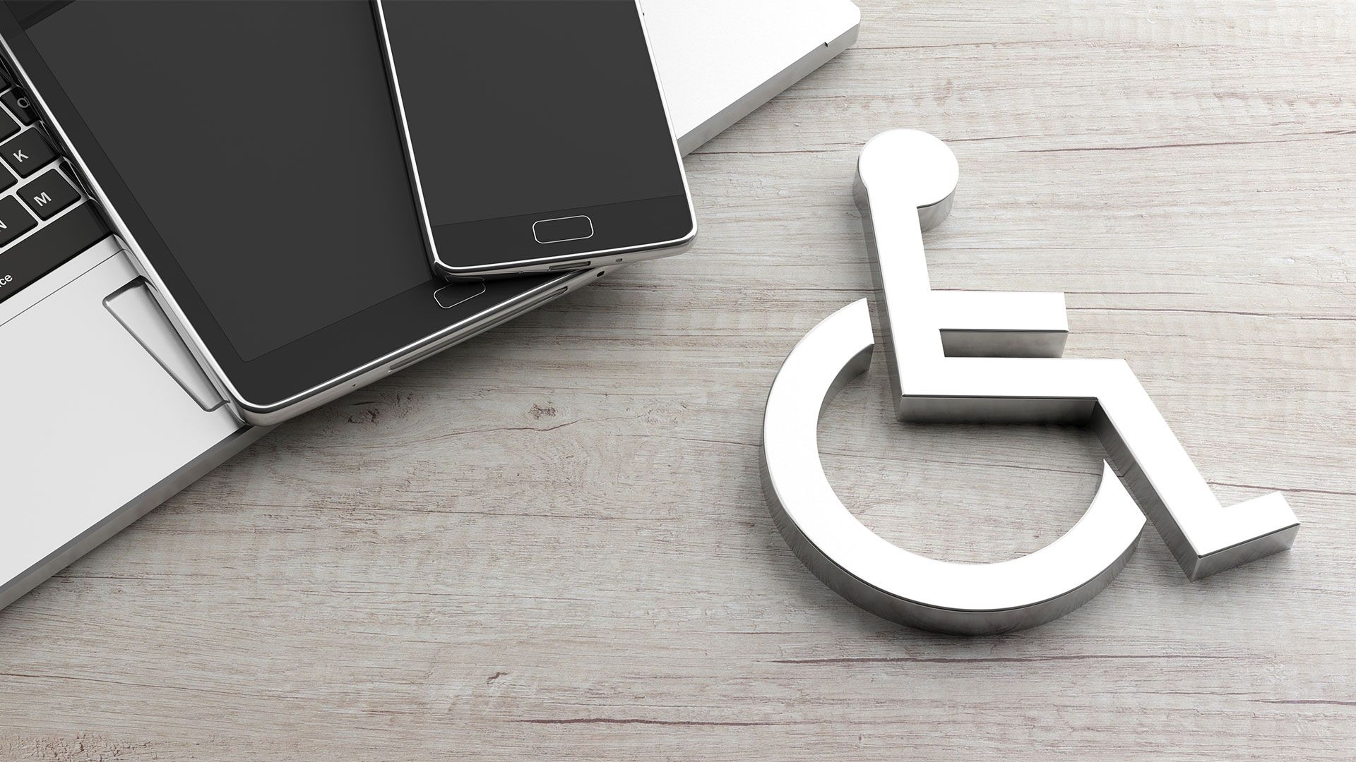 Silver accessibility icon on a wooden desk beside a laptop, tablet, and smartphone.
